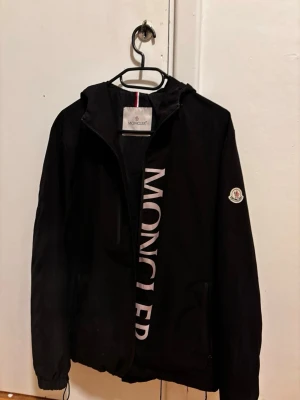 Svart vindjacka från Moncler - Svart vindjacka från Moncler med stor vit logotyp längs dragkedjan och klassisk Moncler-patch på ärmen. Jackan har huva, dragsko nertill och två sidofickor med dragkedja. Perfekt för dig som gillar stilrena och sportiga plagg.