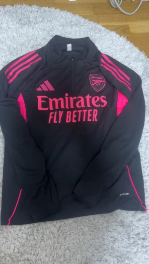Arsenal x Adidas svart och rosa träningsset - Säljer ett Arsenal x Adidas träningsset i svart med starka rosa detaljer. Setet består av långärmad tröja med halv dragkedja och matchande byxor, båda med Arsenal-logga och Adidas stripes. Materialet är syntet och designen är sportig och modern.