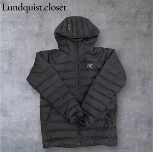 Grå dunjacka från Arc'teryx med huva - Säljer en svart dunjacka från Arc'teryx med huva och dragkedja framtill. Jackan har ett stilrent, puffigt utseende och är perfekt för kalla dagar. Storlek M men passar också väldigt bra som storlek S