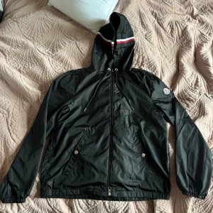 Svart vindjacka från Moncler - Svart vindjacka från Moncler med huva och dragkedja. Jackan har vita och röda ränder på huvan, två fickor med tryckknappar och Moncler-logga på ärmen. Tillverkad i lätt polyester, perfekt för blåsiga dagar. Sitter perfekt på nån som är runt 170. Snygg och Grisch!