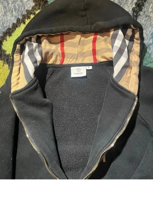 Svart hoodie från Burberry med dragkedja - Säljer en svart hoodie från Burberry med dragkedja och klassiskt rutigt foder i huvan. Hoodien har fickor framtill, ribbade muddar och snörning i huvan. Materialet är mjukt och tjockt, perfekt för kyliga dagar. Stilsäker detalj med Burberrys signaturmönster i huvan.
