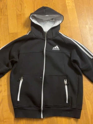 Svart Adidas hoodie med dragkedja - Svart hoodie från Adidas med vit dragkedja, vita detaljer och klassiska ränder längs ärmarna. Adidas-logga på bröstet och ärmen. Två fickor med dragkedja och huva med vit insida. Perfekt för en sportig look.