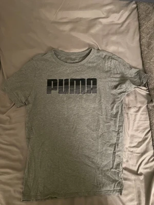 Grå t-shirt från Puma - Snygg grå t-shirt från Puma med svart logga framtill. Klassisk rund halsringning och korta ärmar. Tillverkad i mjuk bomull som är skön att ha på sig. Perfekt för en avslappnad och sportig stil.