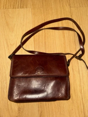 Brun axelväska i skinn från The Trend  - Snygg mörkbrun axelväska i skinn från The Trend. Klassisk rektangulär form med justerbar axelrem och lock med präglad logga. Baksidan har en praktisk ficka med dragkedja. Perfekt för dig som gillar stilrena accessoarer.