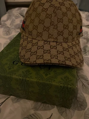 Ingen väst – Gucci keps med GG-mönster - Det här är en beige Gucci keps med klassiskt GG-mönster och grönt/rött band på sidan. Kepsen är tillverkad i bomull och polyester och har justerbart spänne bak. Säljes med originalkartong och dustbag