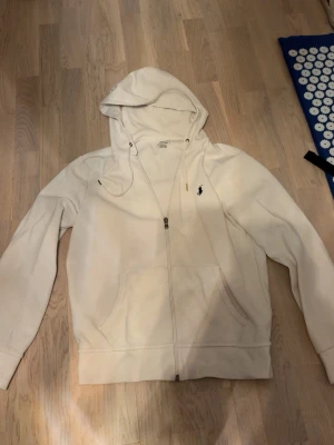 Vit hoodie från Polo Ralph Lauren - Snygg vit hoodie med dragkedja från Polo Ralph Lauren. Klassisk design med huva, två fickor framtill och den ikoniska lilla broderade loggan på bröstet. Perfekt för en avslappnad och stilren look. Tillverkad i mjukt material för extra komfort.