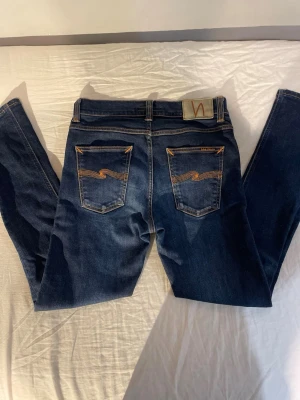 Mörkblå Nudie jeans - Säljer ett par mörkblå jeans från Nudie Jeans med klassiska kontrastsömmar och broderade bakfickor. Jeansen har fem fickor, normal passform och är tillverkade i mjukt bomullsjeansmaterial. Perfekta för dig som gillar en tidlös och clean look.