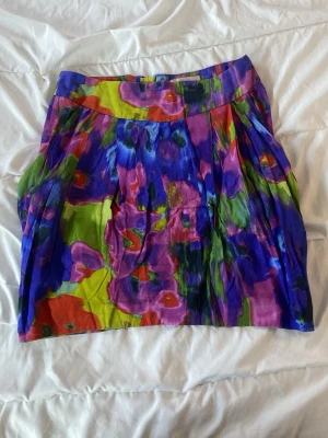 Färgglad blommig kortkjol - Säljer en kortkjol med färgstarkt blommigt mönster i lila, blått, grönt, rött och gult. Kjolen har en bred linning och dragkedja bak. Materialet känns lätt och passar perfekt för varmare dagar. Perfekt statement-plagg för dig som gillar färg!