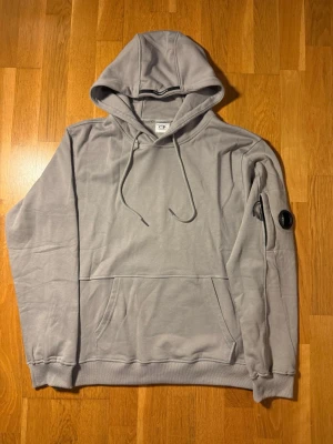 Grå hoodie från C.P. Company - Snygg grå hoodie från C.P. Company med klassisk huva och dragsko. Har en stor magficka och en unik detalj med lins på ena ärmen.