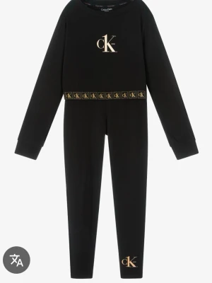 Svart loungewear-set från Calvin Klein - Svart set från Calvin Klein med långärmad topp och matchande byxor. Setet har guldfärgade 'ck' loggor på bröstet, midjan och benet samt elastisk midja med logoband. Tillverkat i mjukt material för en bekväm känsla.