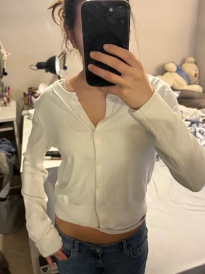 Vit kofta från H&M med knappar - Säljer en klassisk vit kofta från H&M med långa ärmar och knäppning framtill. Koftan är i mjukt material och har en enkel, stilren design som passar till det mesta. Perfekt att slänga över en topp eller t-shirt.