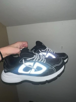 Dior b30 - Snygga svarta Dior sneakers med vita och reflekterande detaljer samt vita snören. Skorna har en tjock vit sula och tydlig Dior-logga på sidan. Perfekta för dig som vill sticka ut med exklusiva och trendiga sneakers.