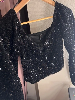 Svart paljettopp  - Glittrig svart croppad topp med långa ärmar och täckt av paljetter. Toppen har en lätt v-ringad halsringning.