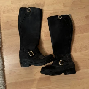 Jonny Bulls stövlar  - Stilrena svarta stövlar med dekorativa spännen och grov sula. Stövlarna har rund tå och dragkedja längs sidan. Perfekta för att lyfta din outfit med en tuff touch.