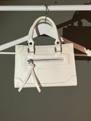 Vit handväska med dragkedja - Stilren vit handväska i syntetmaterial med två handtag och en silverfärgad dragkedja framtill. Väskan har en rektangulär form och minimalistisk design, perfekt för att lyfta din outfit. Snygga sömdetaljer och enkel att matcha. Köpt från Loppis, är i väldigt bra skick 🤍