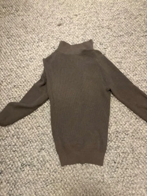 Brun stickad ziptröja från H&M - Säljer en brun stickad tröja från H&M med dragkedja framtill och hög krage. Tröjan har ribbstickad struktur och långa ärmar, perfekt för lager på lager. Enkel och stilren design som funkar till det mesta.