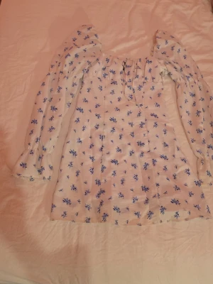 Vit blommig blus med puffärm - Söt vit blus med blått blommönster och puffiga långärmar. Blusen har en fyrkantig ringning och knytning framtill. Tillverkad i ett lätt tyg som ger en romantisk känsla. Perfekt för dig som gillar vintageinspirerad stil.