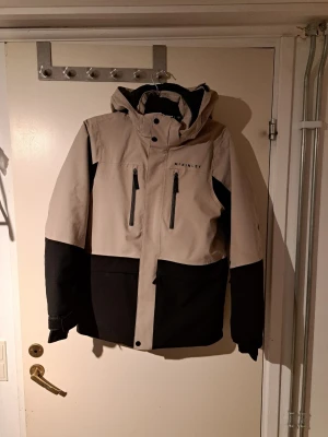 Beige och svart skidjacka McKinley, strl 170 - Snygg beige och svart vindjacka från McKinley med huva och två bröstfickor och två sidofickor med dragkedja. Jackan har hög krage, långa ärmar. Snölås i ärmar och midja och en sportig look. 