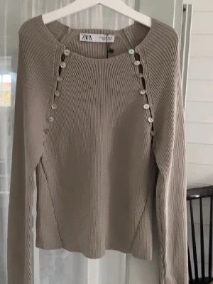 Beige topp från Zara - Snygg beige topp från Zara med rund halsringning och dekorativa knappar längs axlarna. Tröjan har lång ärm och är ribbstickad för en stilren look. Perfekt att matcha med jeans eller kjol. Lånade bilder💕