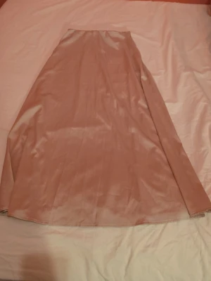 Beige långkjol i satin - Säljer en beige långkjol i mjukt satinliknande material. Kjolen har en enkel och stilren design med lätt glansig yta och rak passform. Perfekt att styla med både sneakers och boots för en trendig look.