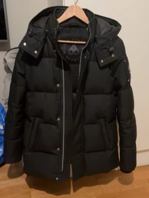 Moose Knuckles - Snygg svart dunjacka från Moose Knuckles med huva och quiltad design. Jackan har dragkedja framtill, tryckknappar och en klassisk pufferlook. Perfekt för kalla vinterdagar och riktigt varm. Logga på ärmen och praktiska fickor.