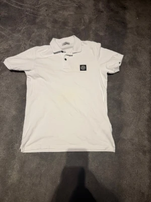 Vit pikétröja från Stone Island Junior - Klassisk vit pikétröja från Stone Island Junior med krage, korta ärmar och knappslå framtill. Tröjan har en liten bröstficka med Stone Island-logga. Perfekt till en clean och stilren look.