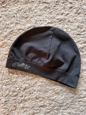 Svart mössa från Sixties  - Snygg svart mössa från Sixties med diskret logga broderad i silver på kanten. Tillverkad i mjukt och stretchigt tyg som sitter skönt på huvudet. Enkel och stilren design som passar till de flesta outfits. Oanvänd helt ny. 