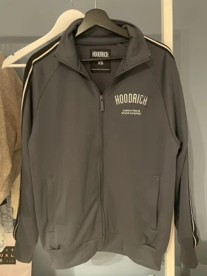Grå ziptröja från Hoodrich - Säljer en grå ziptröja från Hoodrich med vit logga och text på bröstet. Tröjan har hög krage, hel dragkedja, fickor framtill och vita ränder längs ärmarna. Tillverkad i mjukt material som känns skönt mot huden. Perfekt för en sportig och avslappnad stil, Den är i xs men känns mer som s.