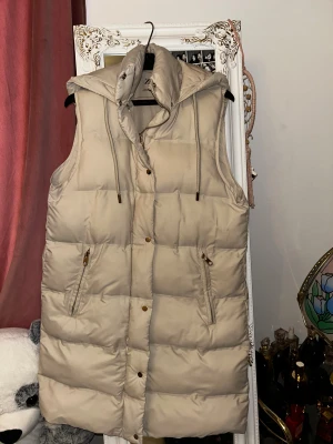 Beige lång dunväst med huva - Säljer en beige lång dunväst med huva och dragsko. Västen har två fickor med dragkedja och stängs med både dragkedja och tryckknappar. Perfekt för lager på lager och riktigt snygg till höst- och vinterlooken.