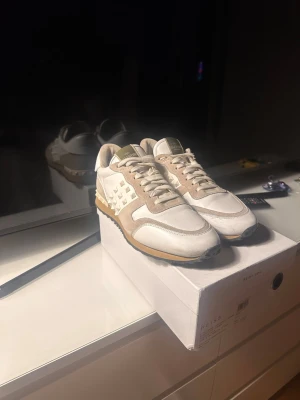 Valentino Garavani Rockrunner sneakers - Snygga Valentino Garavani Rockrunner sneakers i vitt och beige med ikoniska nitar på sidorna och hälen. Skorna har en mix av skinn och mocka, samt klassisk snörning och platt sula. Perfekta för dig som vill sticka ut med stilrena detaljer. INGET OG INGÅR