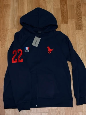 Mörkblå hoodie med tryck Paris 22 - Snygg mörkblå hoodie med dragkedja från Le Coq Sportif. Har röd tupp-logga på bröstet, siffran 22 i rött på ärmen och vitt på ryggen samt Paris-flagga på bröstet. Mjuk bomullskänsla och huva för extra stilpoäng.