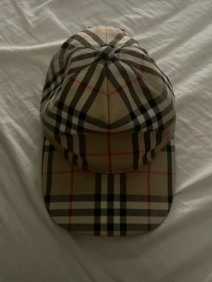 Burberry klassisk rutig keps - Säljer en ikonisk keps från Burberry med det klassiska beigea rutmönstret och justerbar rem bak. Märket har en svart patch med texten 'Burberry London England' på baksidan. Perfekt accessoar för dig som gillar stilrena och tidlösa detaljer.