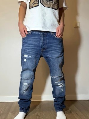 Blå  jeans med slitningar - Säljer ett par blå baggy jeans med slitna detaljer och hål på framsidan. Jeansen har normal midja, raka ben och klassisk femficksdesign. Perfekta för en avslappnad och trendig streetwear-look.