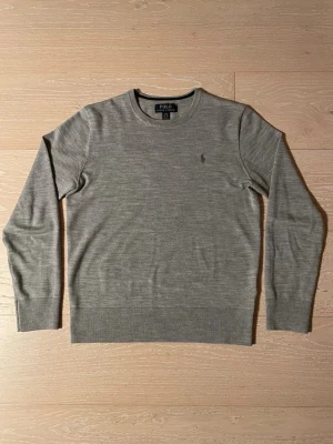 Grå ulltröja från Polo Ralph Lauren - Klassisk grå stickad tröja från Polo Ralph Lauren med rund halsringning och diskret broderad logga på bröstet. Tröjan är i 100% ull och har ribbade muddar vid ärmslut och nederkant. Perfekt för lager-på-lager under dessa kyliga dagar. Tveka inte på att ställa frågor! Pris ej hugget i sten