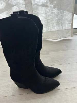 Svarta cowboy boots i mocka - Snygga svarta cowboy boots med spetsig tå och hög klack som ändast är använda fåtal gånger. Tillverkade i mocka med klassiska broderade detaljer på skaftet. Såklart har jag även tvättat skorna noga!!