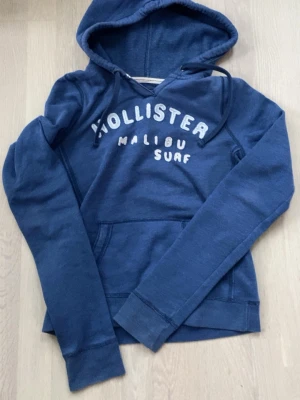 Blå Hollister hoodie med tryck (M fits S) - Snygg blå hoodie från Hollister med broderat vitt tryck 'Malibu Surf' på bröstet. Klassisk huva med dragsko och magficka framtill. Tillverkad i mjukt bomullsmaterial som är perfekt för chill dagar. Långärmad och avslappnad passform.