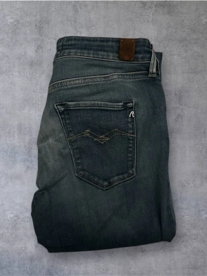 Replay Anbass jeans i mörkblå denim - Snygga Replay Anbass jeans i mörkblå tvätt med klassisk femficksdesign och diskreta slitningar. Jeansen har smal passform och är tillverkade i mjuk bomullsdenim med stretch för extra komfort. Perfekta för en trendig och avslappnad look.