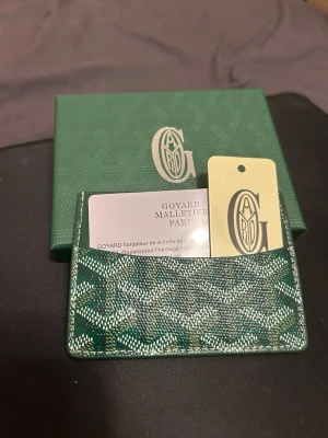 Grönt skinn korthållare  - Kompakt korthållare i grön signaturcanvas med läderdetaljer. Elegant, slitstark och perfekt för kort i ett smidigt format.