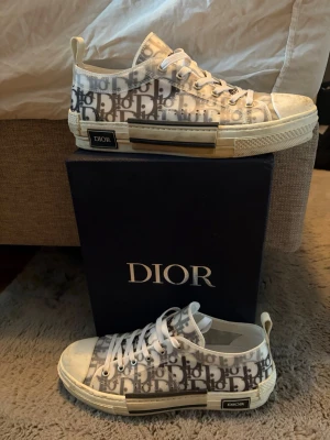 Dior B23 sneakers skor - Säljer ett par Dior B23 sneakers i vitt och grått med det ikoniska Dior-monogrammönstret. Riktigt snygga och trendiga sneakers för dig som gillar lyx. Skorna är storlek 45 men passar också 44. Kvitto medföljer ej men box och dustbag medföljer.