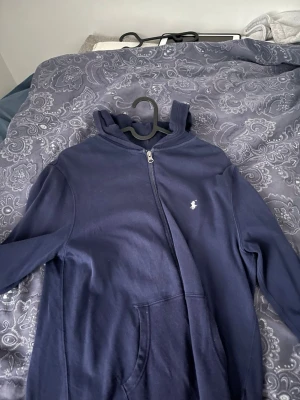 Mörkblå hoodie från Polo Ralph Lauren - Mörkblå hoodie med dragkedja från Polo Ralph Lauren. Klassisk design med huva, fickor framtill och den ikoniska lilla broderade loggan på bröstet. Tillverkad i mjukt bomullsmaterial som är skönt att ha på sig. Perfekt för en avslappnad och stilren look.
