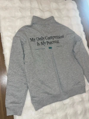 Grå halvzip hoodie med tryck - Grå hoodie med halv dragkedja och hög krage. Baksidan har texten 'My Only Competition Is My Potential.' Perfekt för en chill och sportig look.