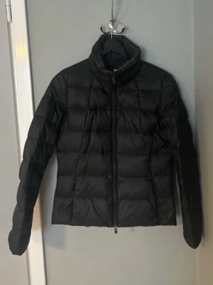 Svart dunjacka från Boss Orange - Svart dunjacka från Boss Orange med quiltad design och hög krage. Jackan har dragkedja framtill och ett mönstrat innerfoder. Perfekt för kyliga dagar och enkel att matcha med olika outfits.