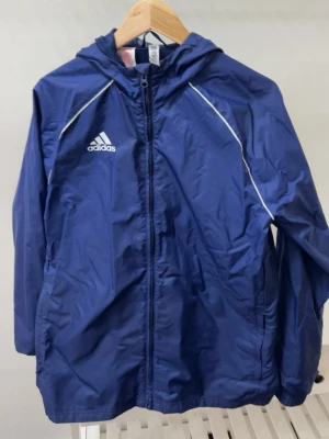 Blå vindjacka från Adidas med huva - Blå vindjacka från Adidas med vit logga på bröstet och vita detaljer längs axlarna. Jackan har huva, dragkedja framtill och är perfekt för blåsiga dagar. Sportig look och klassisk Adidas-stil.