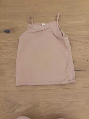 Beige linnetopp med smala axelband - Säljer en enkel och stilren beige linnetopp från Åhléns med smala axelband. Toppen är ärmlös och har en liten rosett framtill vid halsringningen. Perfekt att matcha med jeans eller kjol för en avslappnad look.