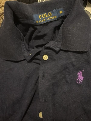 Mörkblå pikétröja från Ralph Lauren - Klassisk mörkblå pikétröja från Ralph Lauren med krage och knappar framtill. Tröjan har det ikoniska lila Polo-logotypbroderiet på bröstet och är tillverkad i mjuk bomull. Perfekt för en stilren och avslappnad look.