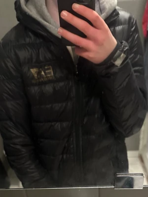 Svart EA7 dunjacka med huva - Snygg svart dunjacka från EA7 Emporio Armani med guldfärgad logga på bröstet. Jackan har huva, dragkedja framtill och quiltad design som ger en modern look. Perfekt för kalla dagar och enkel att matcha med olika outfits.