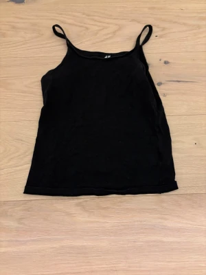 Svart ribbad crop top från H&M - En enkel svart crop top från H&M med tunna axelband och rak halsringning. Tillverkad i mjuk bomull som känns skön mot huden. Perfekt att matcha med jeans eller kjol för en clean och stilren look.