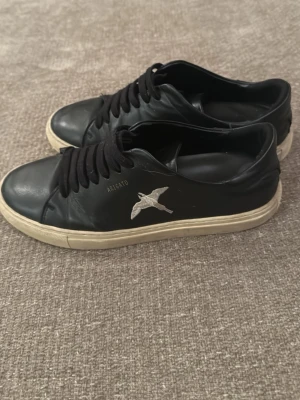 Svarta Axel Arigato sneakers i skinn - Svarta sneakers från Axel Arigato med vit broderad fågel på sidan. Skorna har snörning och är tillverkade i skinn, perfekt för en stilren och modern look. 