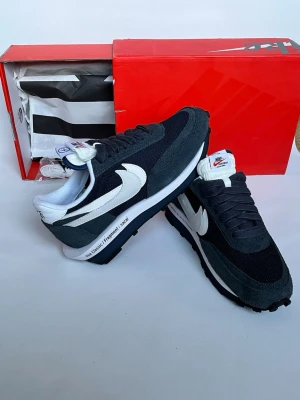 Nike LDWaffle x Sacai x Fragment svart/vit - Nike LDWaffle i samarbete med Sacai och Fragment. Svarta och mörkgrå sneakers med dubbla vita swooshes, mesh och mocka i ovandelen samt vit mellansula med textdetaljer. Klassisk retrostil med svart yttersula och dubbla tungor.