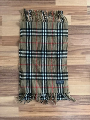 Burberry halsduk - Klassisk halsduk i beige med svart, vitt och rött rutmönster från Burberry. Halsduken har fransar i ändarna och är tillverkad i mjukt material. Perfekt accessoar för att lyfta din outfit.